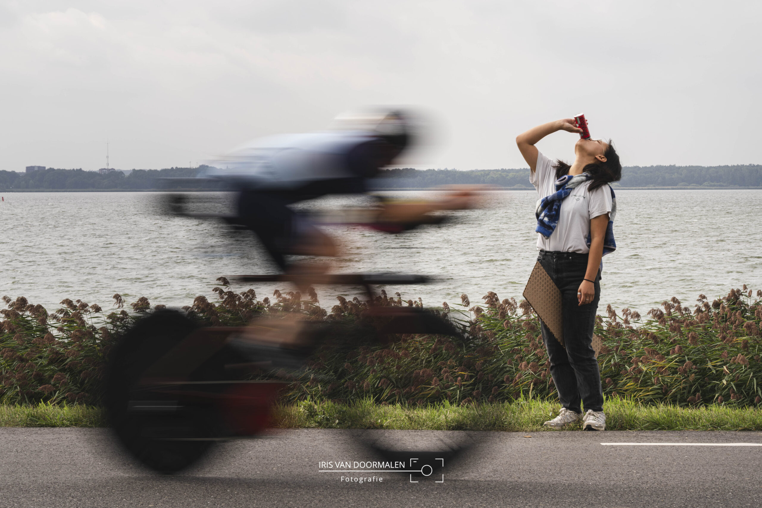 triatlon almere, toeschouwer heeft aanmoedigingsbord tussen haar benen en drinkt haar blikje cola leeg, terwijl triatleet op fiets voorbij raast